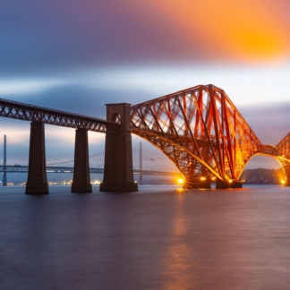 A tranquil evening view of the Forth Bridge, a railway bridge spanning the Firth of Forth near Queensferry, its illuminated red lattice structure reflected in the calm waters.